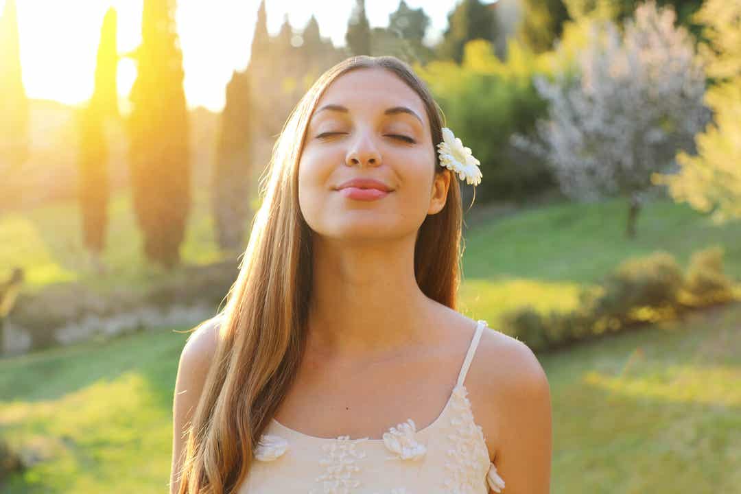 A woman smiling with her eyes closed while standing in the middle of nature.