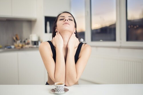 A woman at a kitchen counter drinking coffee and stretching her neck to relieve her neck pain.