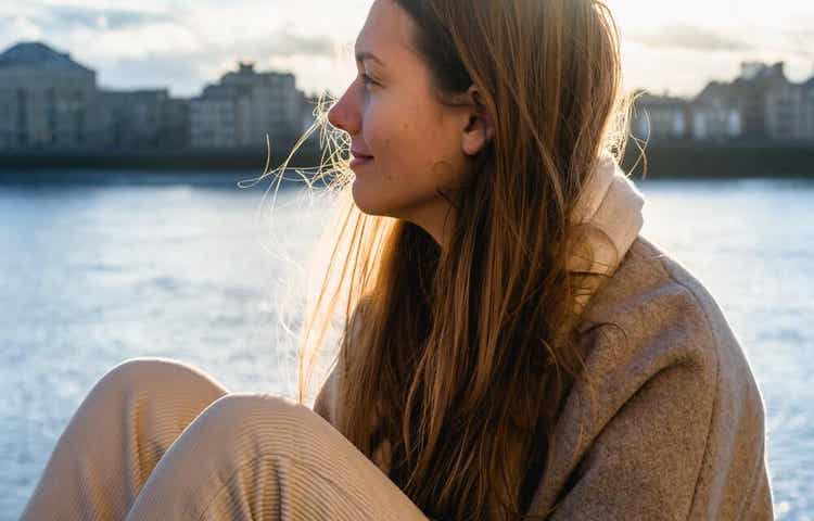 A woman sitting on the edge of a river, smiling thoughtfully.