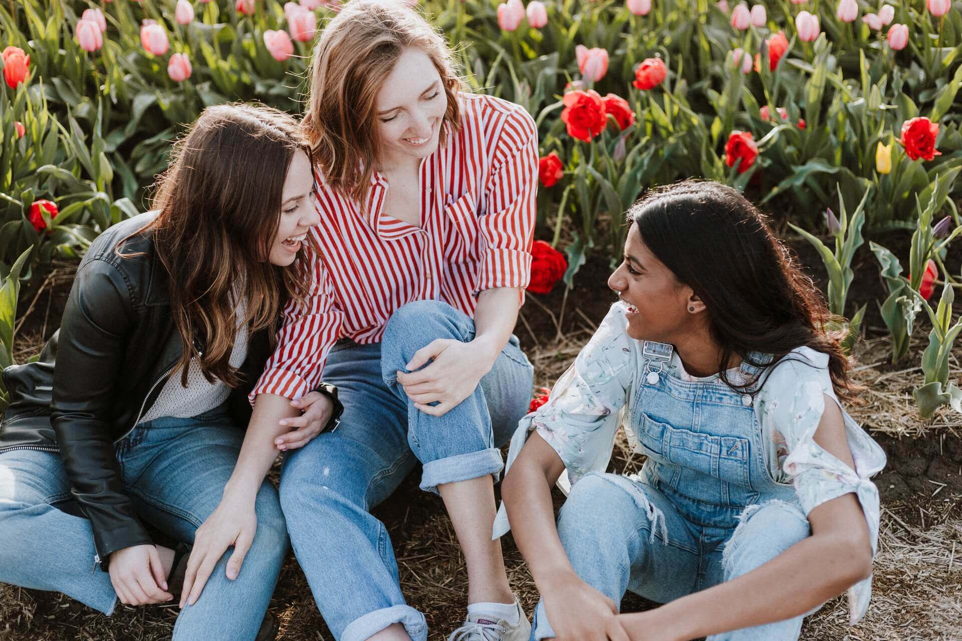Three young women sitting on the ground talking and smilking, with a field of tulips behind them.