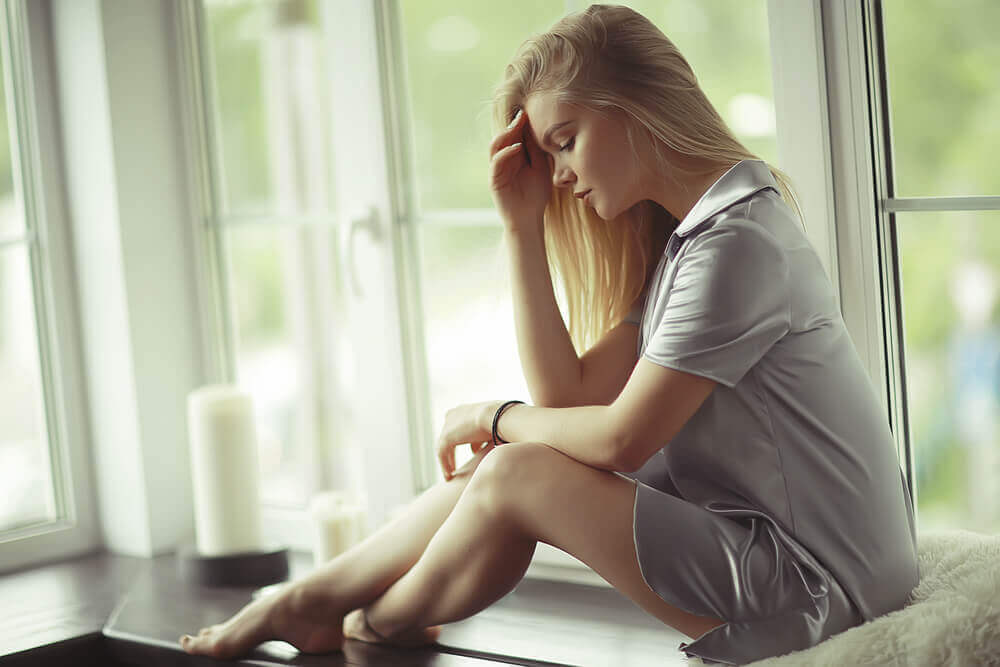 Woman with her hand to her head, sitting on a window sill going through a difficult situation
