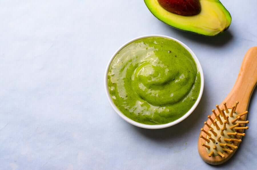 A small bowl of avocado hair mask next to a wooden hair brush and a avocado half.