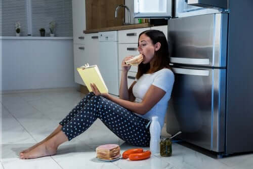 A woman reading and eating by the fridge.