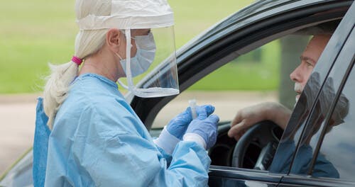 Health professional wearing PPE standing by car to test patient.