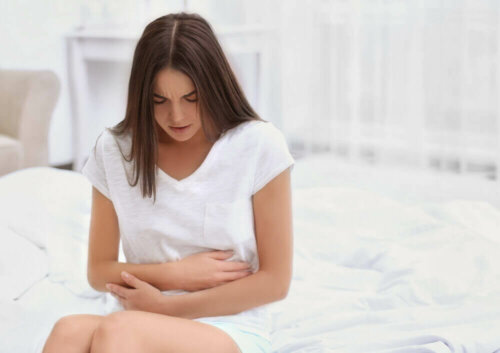 Woman in white sitting on side of bed holding stomach in pain.