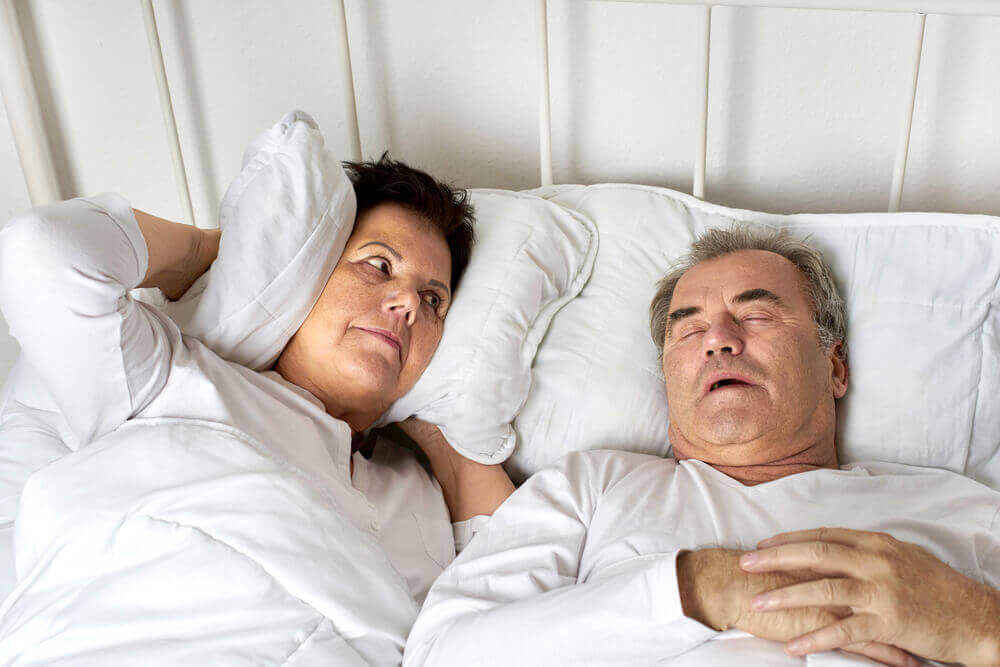 A man snoring in bed while his wife covers her ears with a pillow.