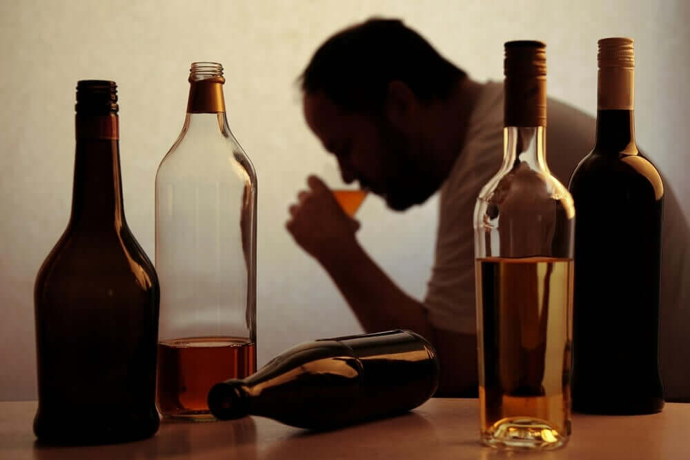 Man drinking alcohol alone in a room with empty bottles around him.