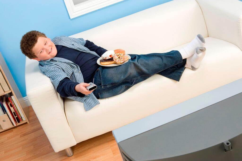 Dziecko z nadwagą jedzące pączki siedząc na kanapie i oglądając telewizję.