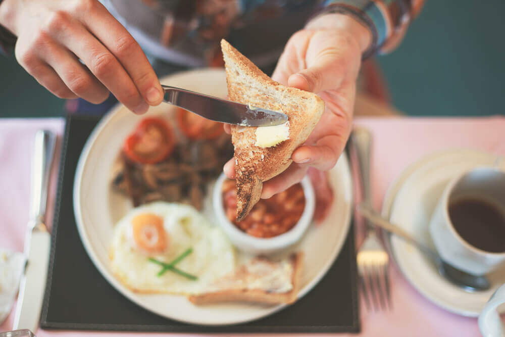 A man spreading butter on toast, with a plate full of breakfast foods in the background.