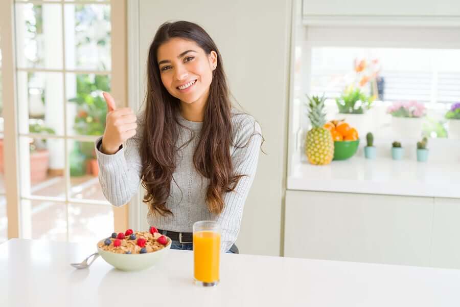 A woman giving a thumbs up as she sits at the counter with a bowl of cereal and a glass of orange juice.