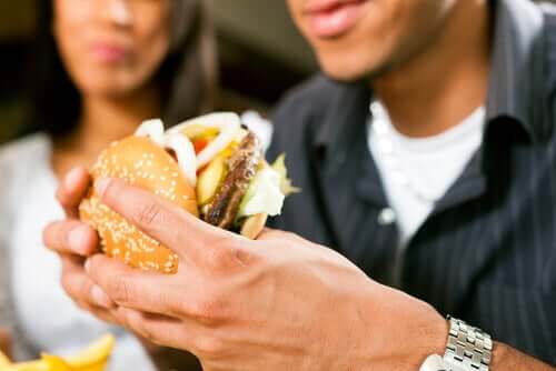 A man eating a double hamburter loaded with condiments as a woman watches in the background.