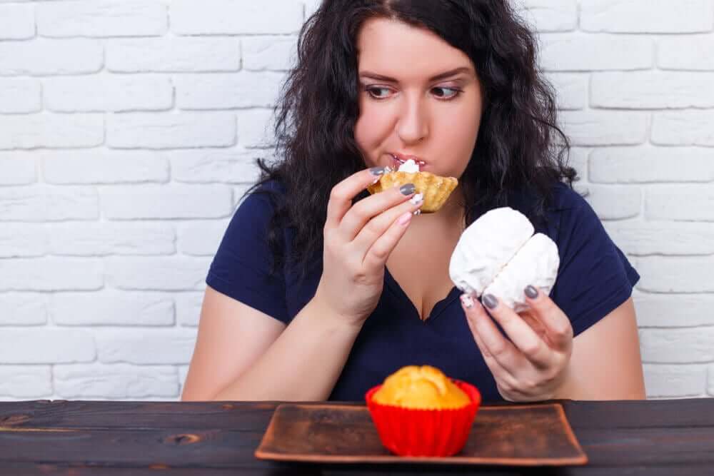 A woman with a different pastry in each hand and a cupcake on the table in front of her.
