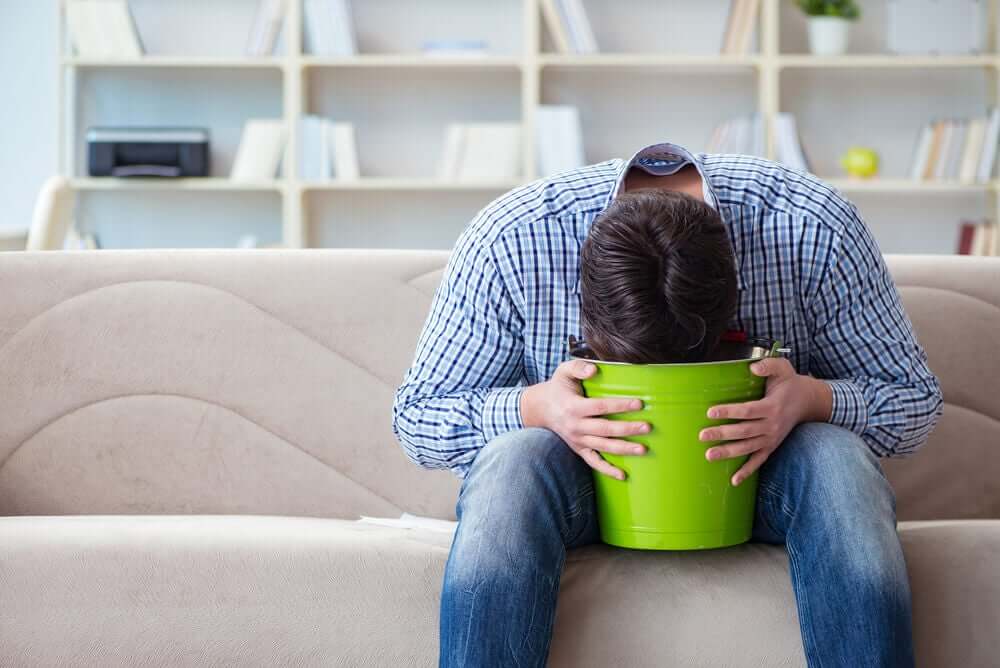 A man sitting on a couch with his head over a bucket because he feels nauseous.