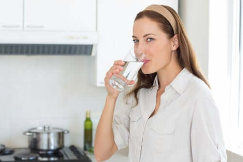 A woman smiling and drinking a glass of water while standing in her kitchen.