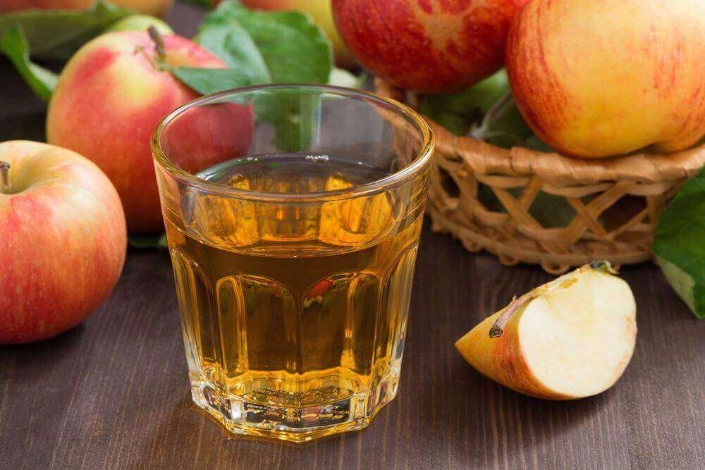 Two glasses of apple juice with whole and cut apples in the background.