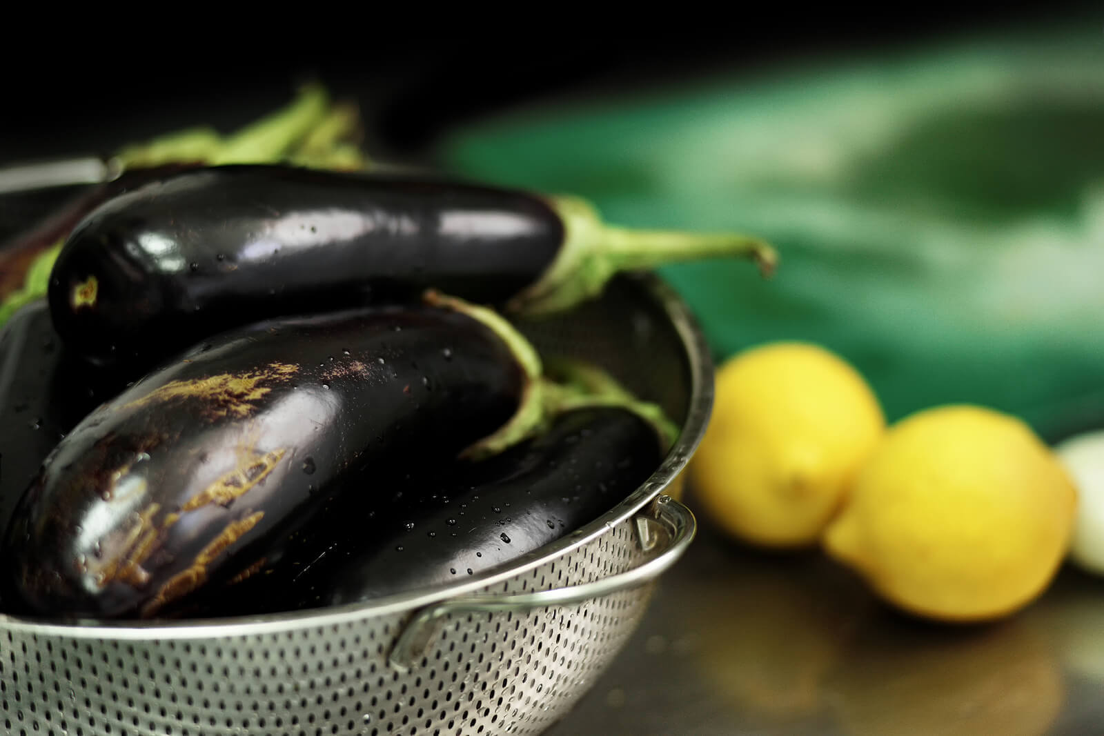 Whole eggplants in a strainer, with whole lemons in the background.