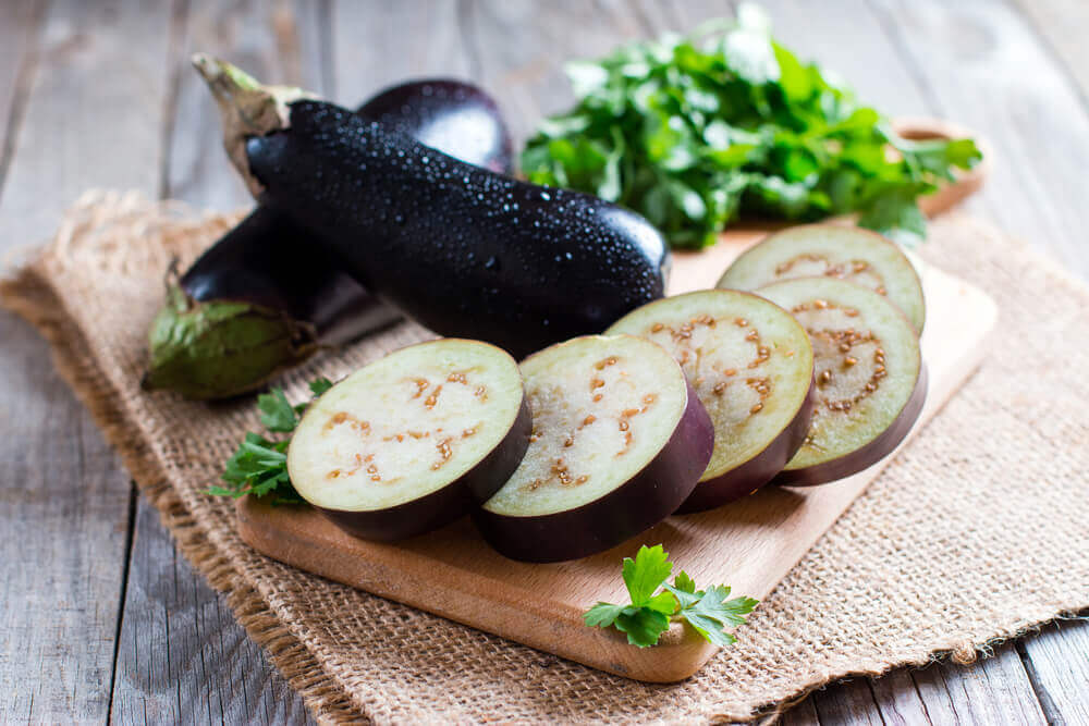 Sliced eggplant on a cutting tray, with whole eggplant and parsley in the background.