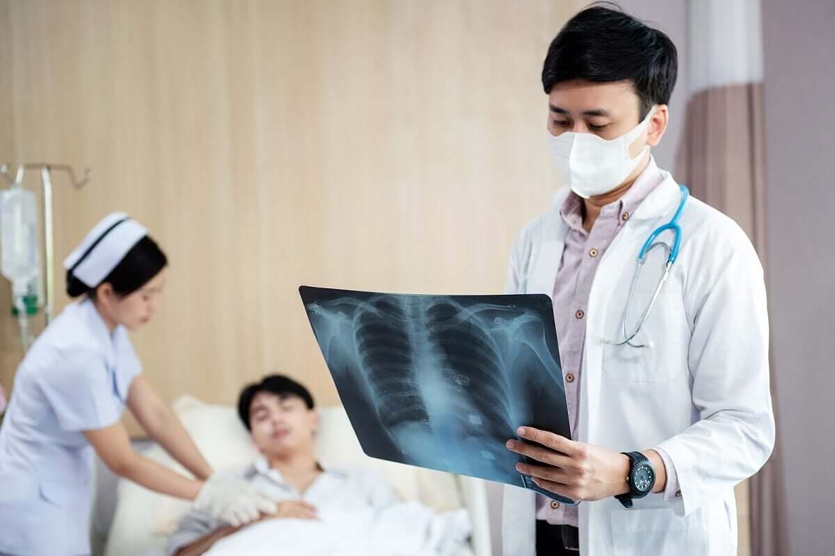 A doctor looking at a lung x-ray with a patient in a hospital bed in the background.