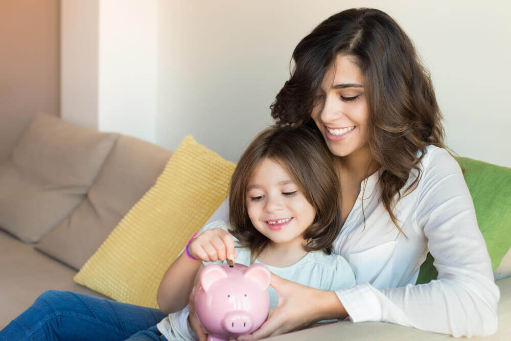 A young girl putting money in a piggy bank while sitting on her mom's lap.