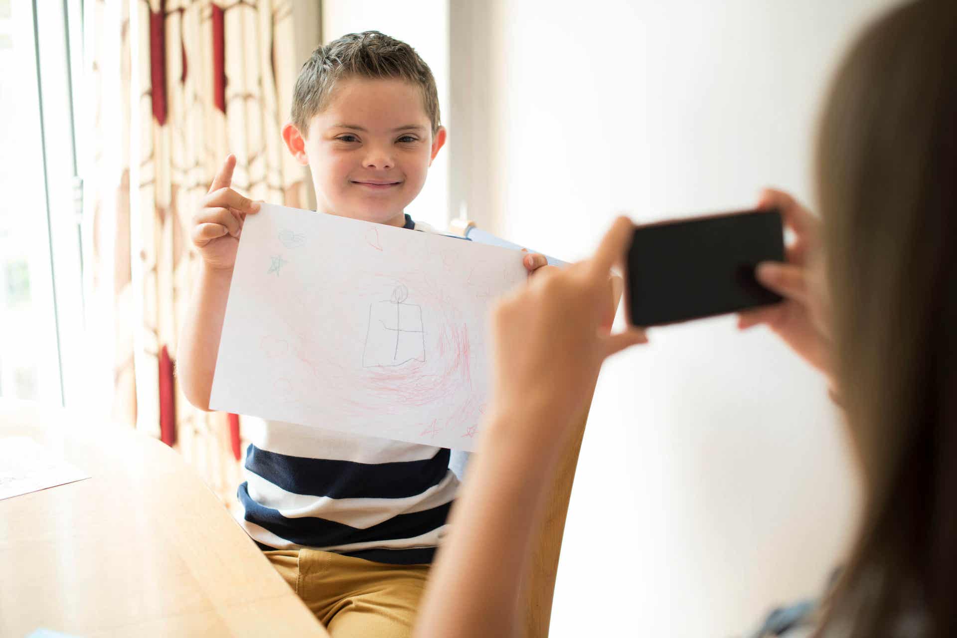 A boy with down syndrome holding up his drawing while his mom takes a picture.